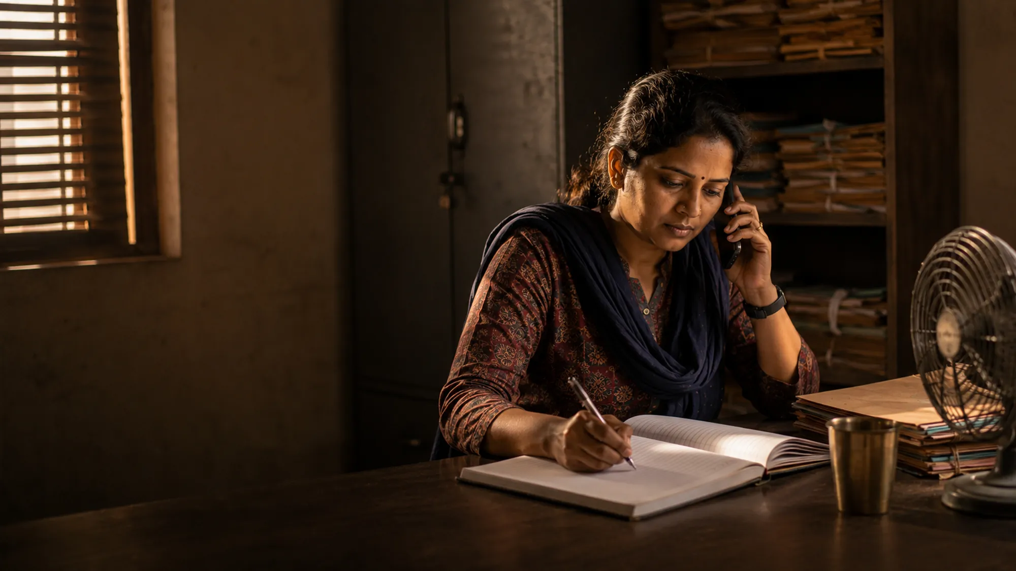 A field coordinator on the phone at a desk in a small office, writing in an open notebook.