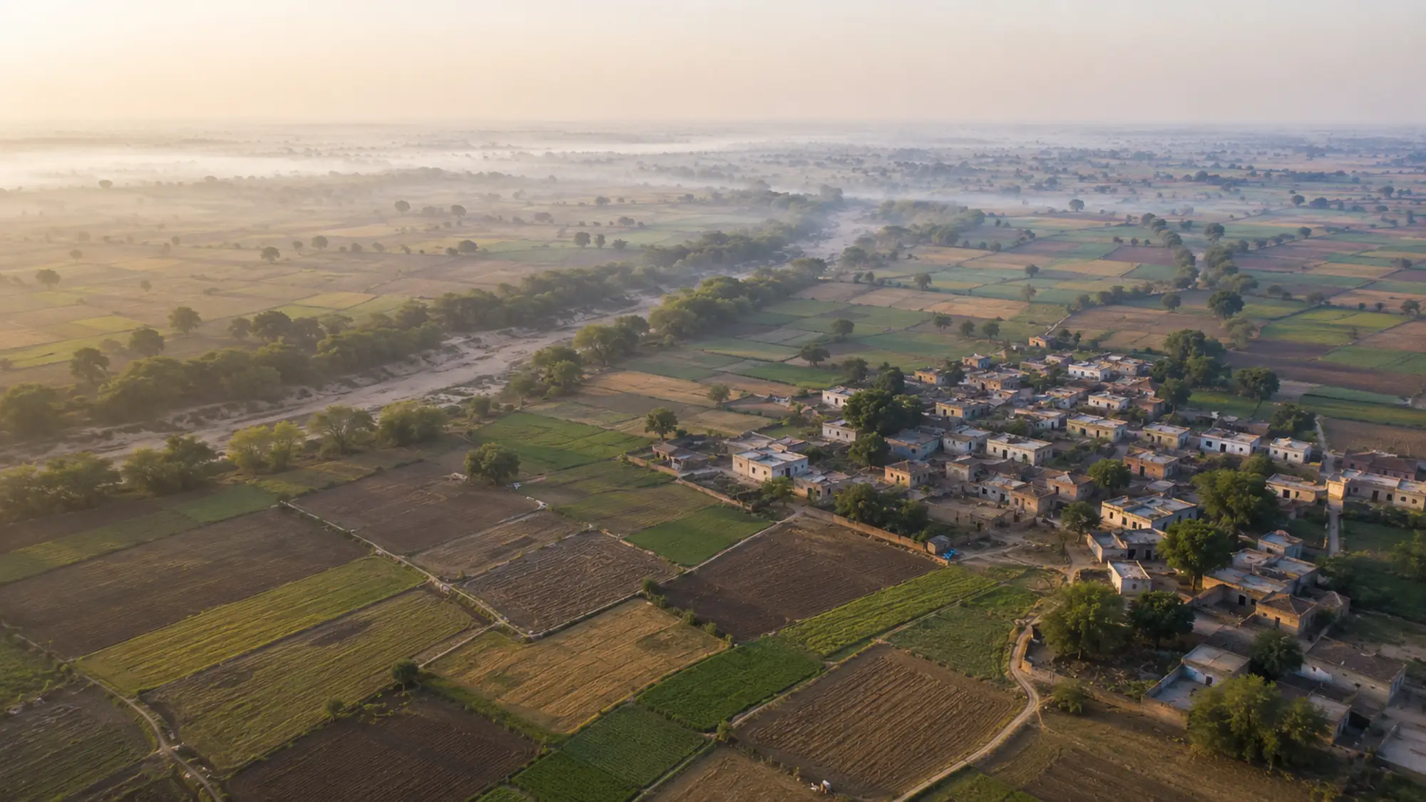Aerial view of a rural Indian village at sunrise, surrounded by a patchwork of small cultivated fields.