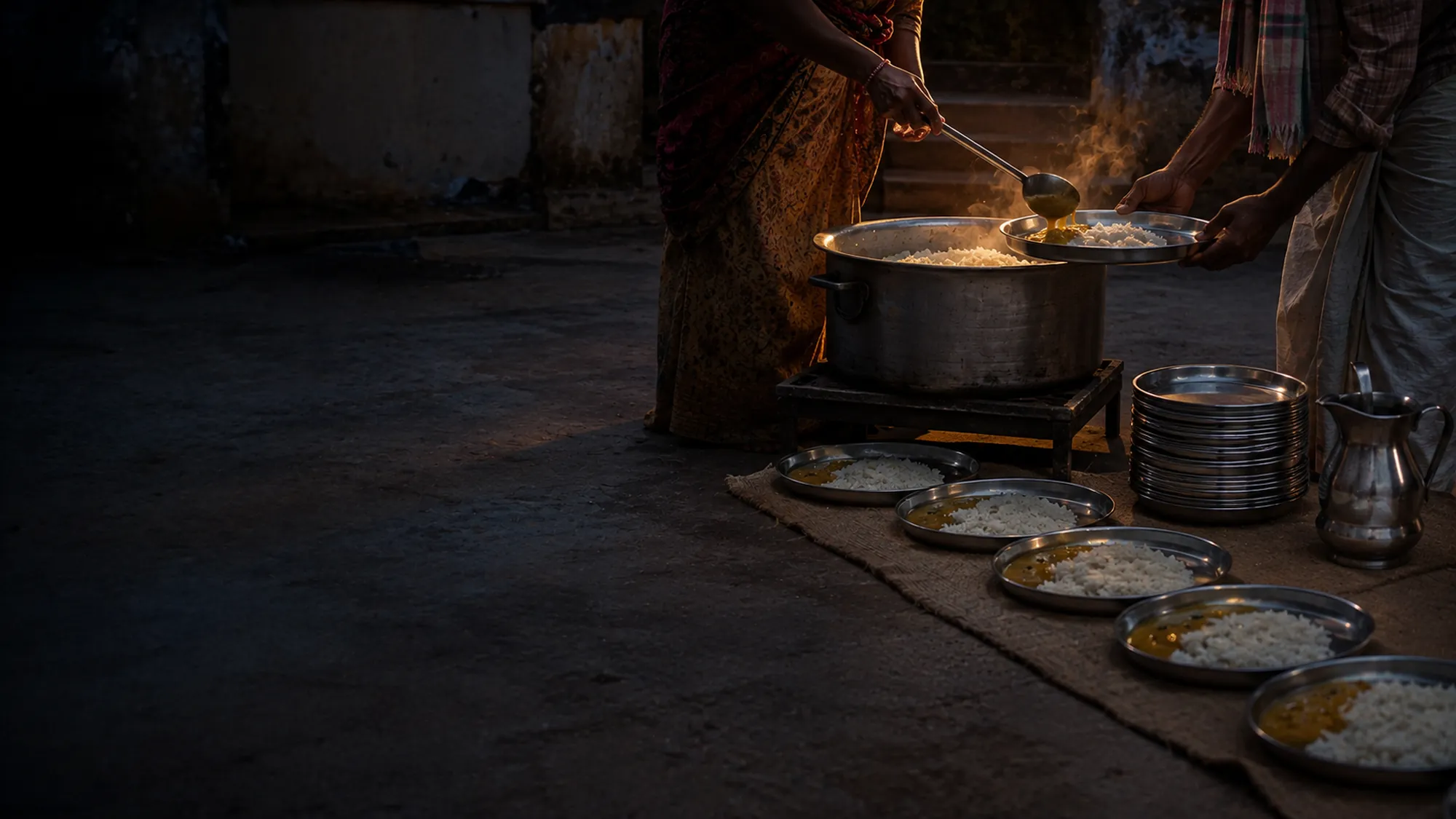 A simple shared meal of rice and dal being served into steel plates at dusk.